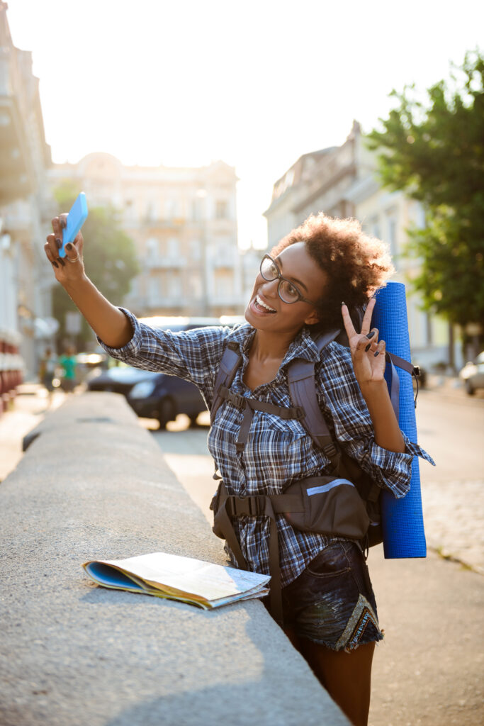 Young beautiful african female traveler with backpack smiling, making selfie.