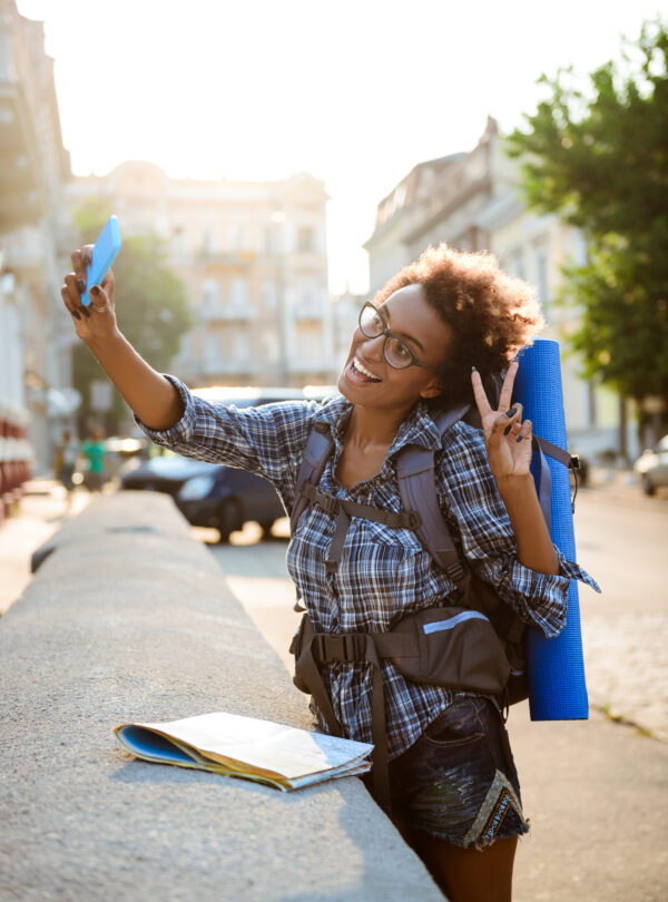 Young beautiful african female traveler with backpack smiling, making selfie.